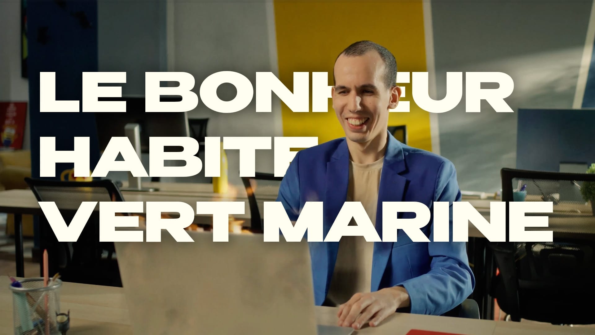 Une personne portant une veste bleue sourit tout en travaillant sur un ordinateur portable à un bureau avec le texte « LE BONHEUR HABITE VERT MARINE » en grosses lettres.